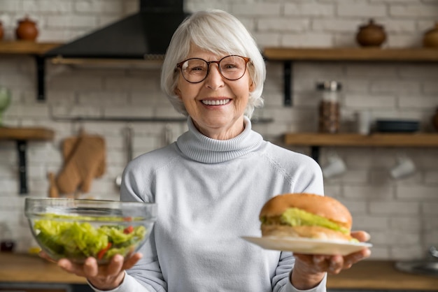 Concepto de comida saludable Mujer sonriente madura sosteniendo ensalada y hamburguesa