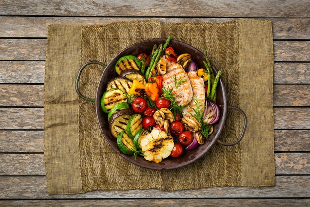 Concepto de comida de verano estacional. Verduras asadas y pechuga de pollo en una sartén sobre una mesa de madera. Vista superior de fondo plano laico