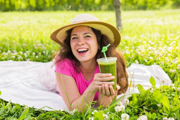 Concepto de comida, verano y personas saludables: la mujer joven se divierte en el parque y bebe batidos verdes en un picnic