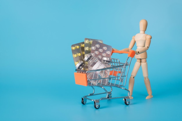 Concepto de compra de drogas. Foto de un hombre de madera que lleva un carrito de compras lleno de diferentes píldoras farmacéuticas aislado fondo azul