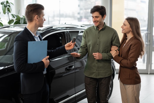 Concepto de compra o arrendamiento de automóviles alegre pareja joven tomando la llave del automóvil del vendedor de automóviles en el moderno