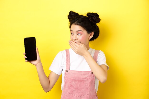 Concepto de compra online. Chica japonesa tonta con maquillaje de belleza, tapa la boca con la mano riendo y mostrando la pantalla vacía del teléfono inteligente, muestra algo gracioso en el teléfono, amarillo.