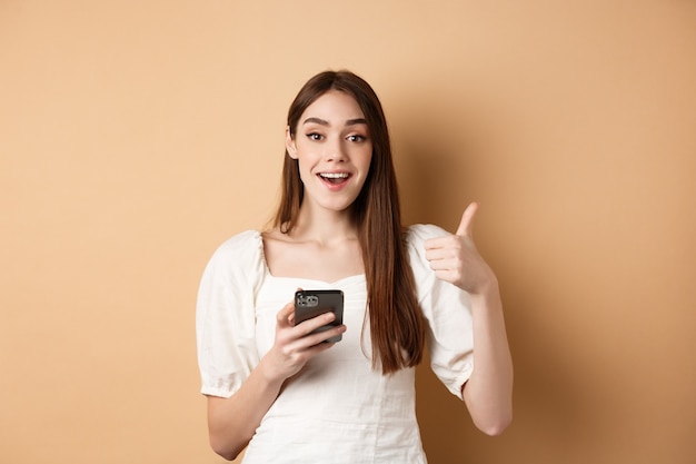 Concepto de compra online. Feliz joven mostrando el pulgar hacia arriba y usando el teléfono celular, sonriendo complacido, de pie sobre fondo beige.