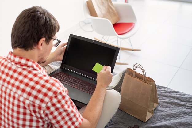 Concepto de compra online. Hombre joven sentado en la cama y compras en línea usando la computadora portátil mirando la tarjeta creadit, pantalla en blanco