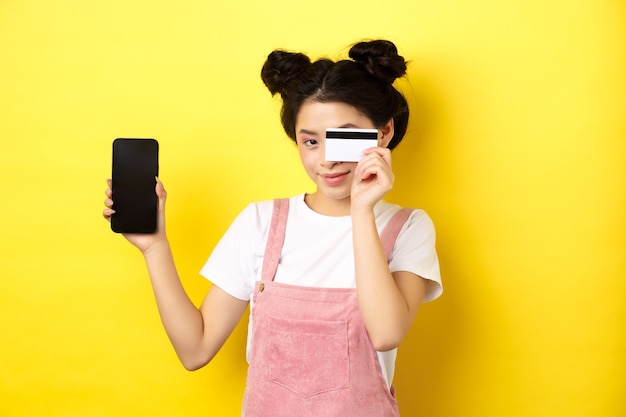 Concepto de compra online. Linda chica asiática pagando con tarjeta de crédito plástica, mostrando la pantalla vacía del teléfono inteligente y sonriendo, amarillo