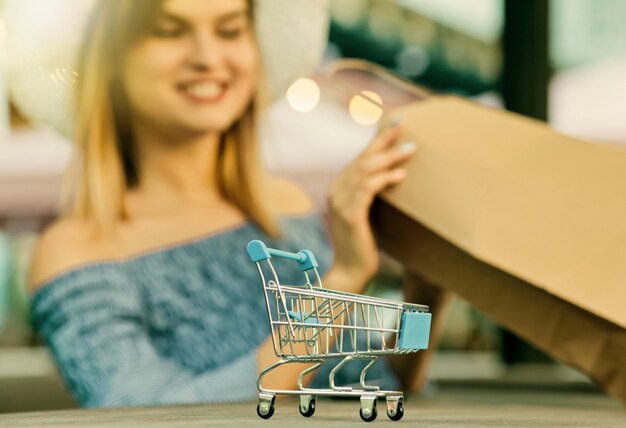 Concepto de compra online. Mini carrito de compras de cerca con fondo borroso de joven atractiva adicta a las compras