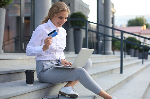 Concepto de compra online. Mujer rubia joven que sostiene una tarjeta de crédito y que hace el pago en línea con la computadora portátil al aire libre.