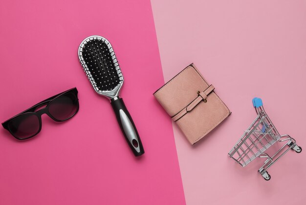 Concepto de compras accesorios para mujer de moda billetera de cuero peine gafas de sol mini carrito de compras sobre fondo de color rosa