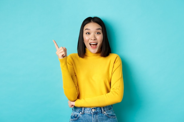 Concepto de compras. Atractiva mujer coreana sonriendo asombrada, apuntando con el dedo hacia la izquierda, mostrando un banner de buen trato, de pie contra el azul.