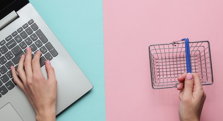 Foto concepto de compras en línea manos femeninas sostienen la mini cesta de la compra y escriben en el teclado del portátil sobre fondo rosa azul pastel