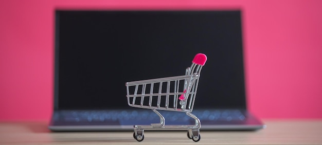 Concepto de compras en línea Mini carro de supermercado y computadora portátil sobre un fondo rosa Carro pequeño y computadora personal