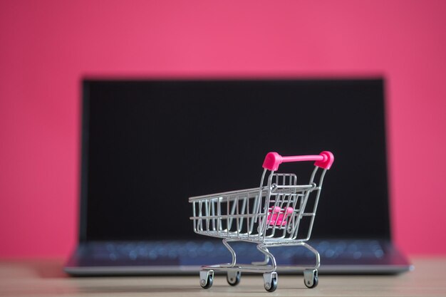 Concepto de compras en línea Mini carro de supermercado y computadora portátil sobre un fondo rosa Carro pequeño y computadora personal