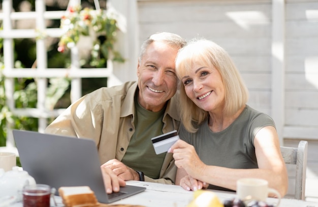 Concepto de compras en línea pareja senior moderna usando computadora portátil y tarjeta de crédito sentado al aire libre en