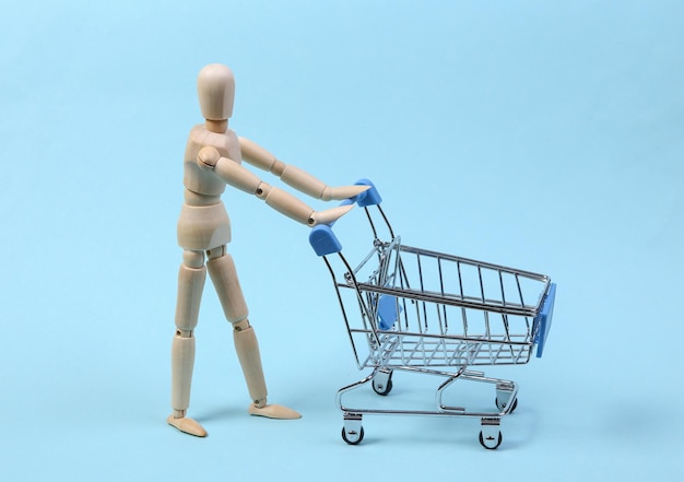 Concepto de compras. Marioneta de madera con carrito de supermercado sobre fondo azul.