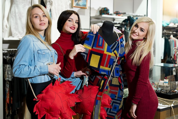 Concepto de compras, moda y amistad - tres amigos sonrientes probándose algo de ropa en el centro comercial.