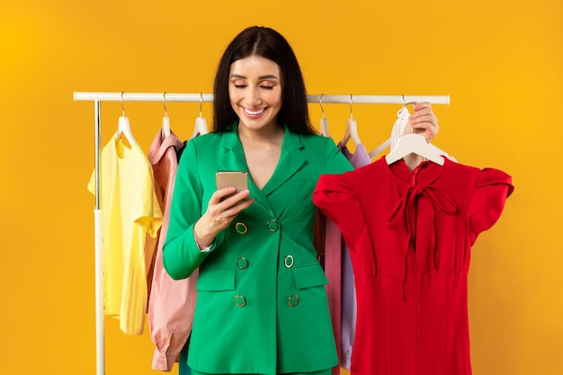 Concepto de compras móviles Señora feliz usando teléfono celular sosteniendo percha con nuevo vestido rojo ordenando tendencias de moda de verano