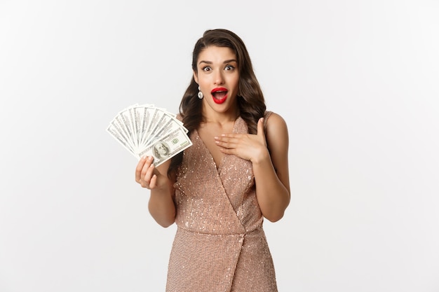 Concepto de compras. Mujer elegante en vestido de glamour con dinero y mirando sorprendido. premio ganador, de pie sobre fondo blanco