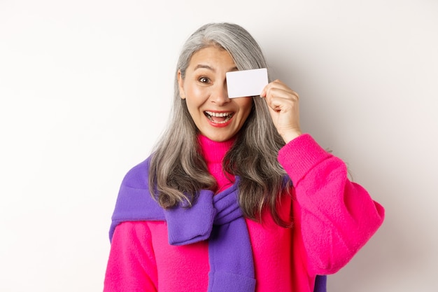 Concepto de compras. Mujer mayor asiática elegante que sonríe y que muestra la tarjeta de crédito plástica, que paga sin contacto, que se coloca sobre el fondo blanco.