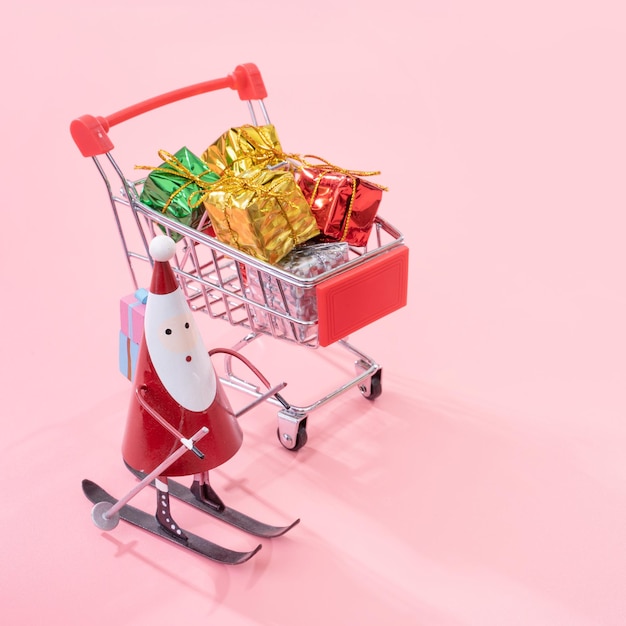 Concepto de compras navideñas mini carrito de compras rojo con juguete de Santa Claus y caja de regalo aislado sobre fondo rosa pálido espacio de copia en blanco cerrar