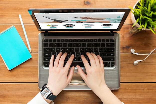 Concepto de compras, negocios, personas y tecnología en línea: cierre de una mujer escribiendo en una computadora portátil con una página web de la tienda de Internet en la pantalla, cuaderno y auriculares en una mesa de madera