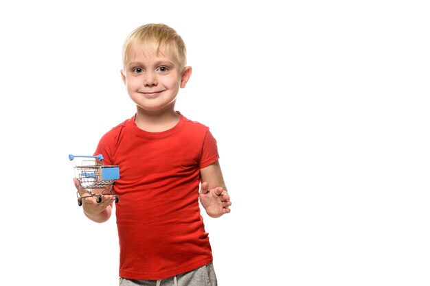 Concepto de compras. Pequeño muchacho sonriente lindo rubio en la camiseta roja que sostiene un pequeño carro de compras del metal. Aislar en blanco