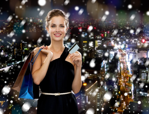 concepto de compras, venta, navidad, gente y vacaciones - mujer sonriente vestida de noche con bolsas de compras sobre el fondo nevado de la ciudad nocturna