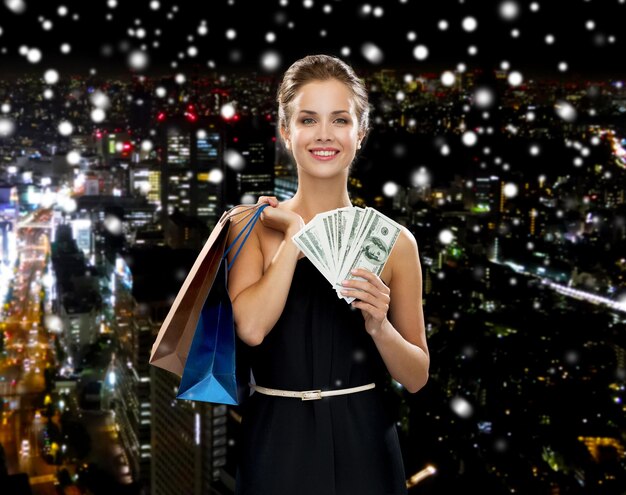 Foto concepto de compras, venta, regalos, dinero y vacaciones - mujer sonriente vestida con bolsas de compras y dinero sobre el fondo de la ciudad de noche nevada