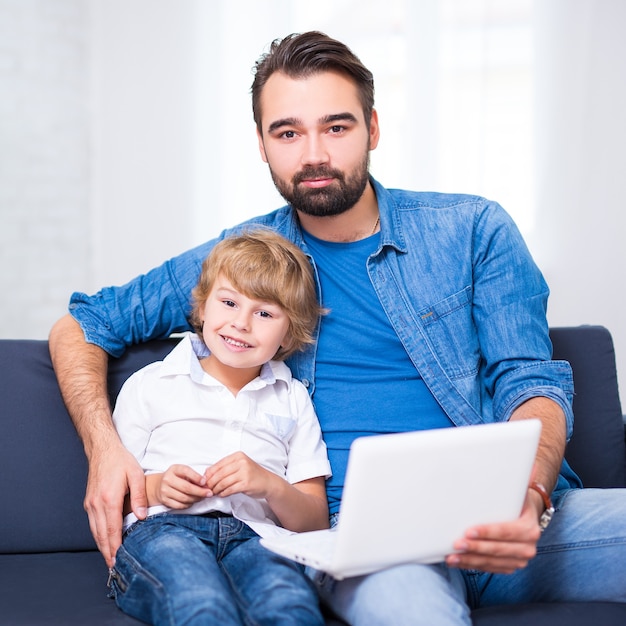 Concepto de computadora e internet - padre joven e hijo pequeño sentado en el sofá con computadora portátil blanca