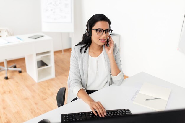 Concepto de comunicación, negocios, personas y tecnología: empresaria sonriente u operadora de línea de ayuda con auriculares y computadora hablando y escribiendo en la oficina