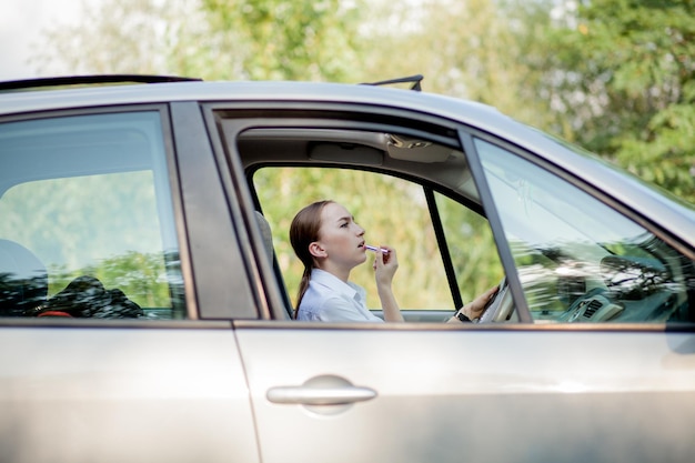 Concepto de conducción peligrosa Joven conductora pelirroja adolescente pintándose los labios maquillándose mientras conduce el coche