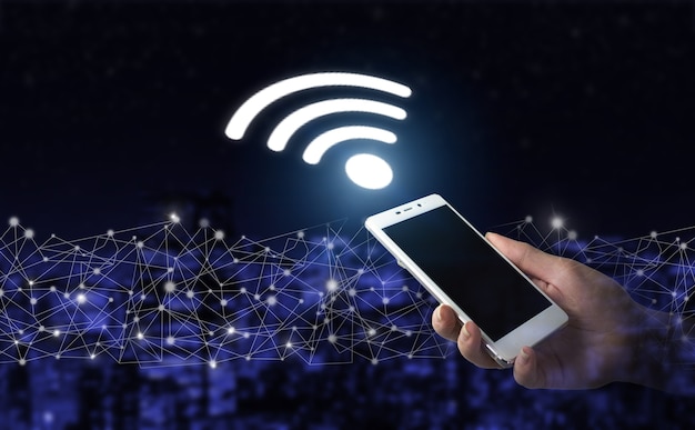 Concepto de conexión de redes empresariales y Wi-Fi en la ciudad. Asimiento de la mano smartphone blanco con señal de Wi Fi holograma digital sobre fondo borroso oscuro de la ciudad. Concepto inalámbrico Wi Fi.