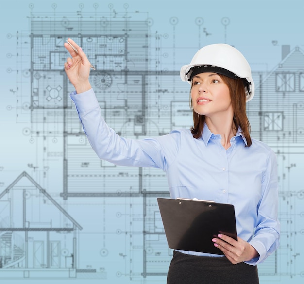 concepto de construcción, desarrollo, construcción y arquitectura - mujer de negocios sonriente con casco blanco y portapapeles apuntando con el dedo