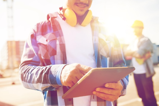 concepto de construcción, tecnología y personas - cierre de constructor sonriente con auriculares y computadora de tablet pc al aire libre