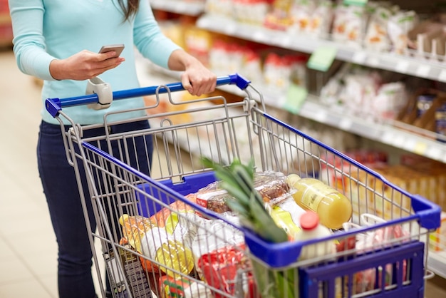 Concepto de consumismo, tecnología y personas: mujer con teléfono inteligente y carrito de compras o carrito comprando comida en una tienda de comestibles o supermercado