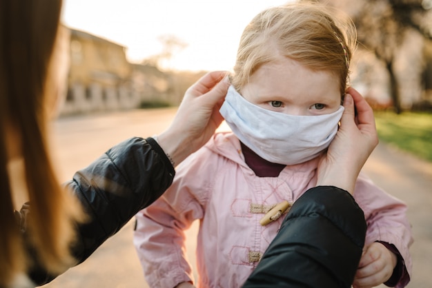 Concepto de coronavirus y contaminación del aire. Niña y madre con máscaras a pie en la calle. Mamá corrige la máscara infantil. Síntomas del virus pandémico. Familia con niños al aire libre. Protección contra enfermedades.