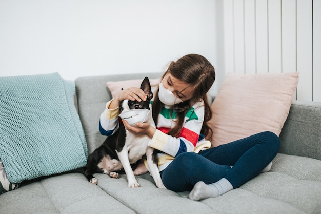 Concepto de coronavirus COVID-19. Niña y su perro con máscaras protectoras en la cara juegan a vivir mientras están en cuarentena.