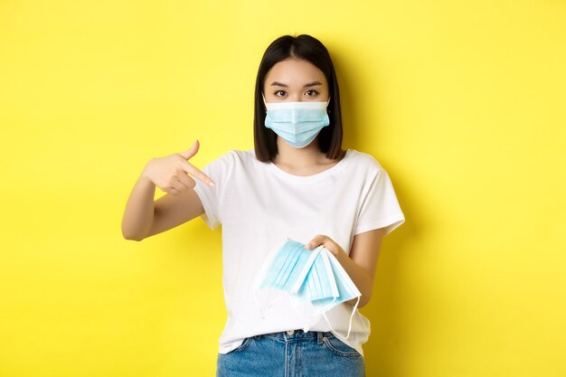 Concepto de coronavirus, cuarentena y medicina. Mujer asiática joven que señala con el dedo a las máscaras médicas para salir al aire libre, de pie sobre fondo amarillo.