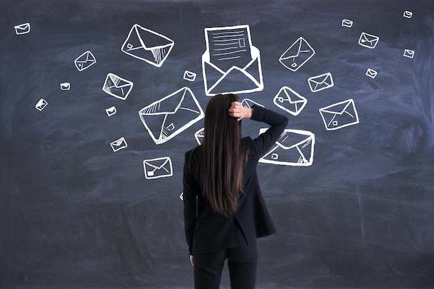 Concepto de correo y marketing con vista posterior de una mujer pensativa con traje negro mirando una pizarra con sobres escritos a mano