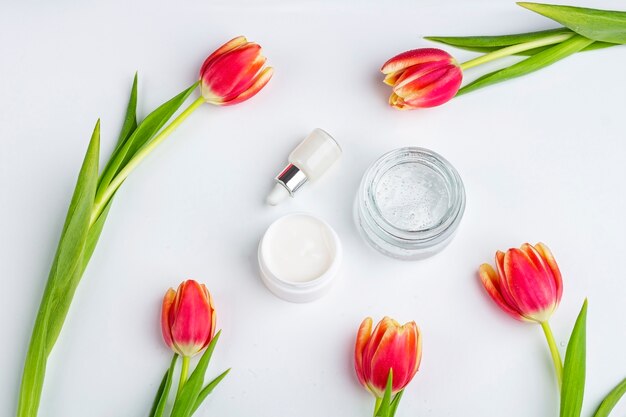 Concepto de cosmética casera orgánica natural. productos para el cuidado de la piel, remedios y belleza: envases con crema y suero entre flores de tulipán rojo primaveral sobre superficie blanca. lay flat, copia espacio para texto