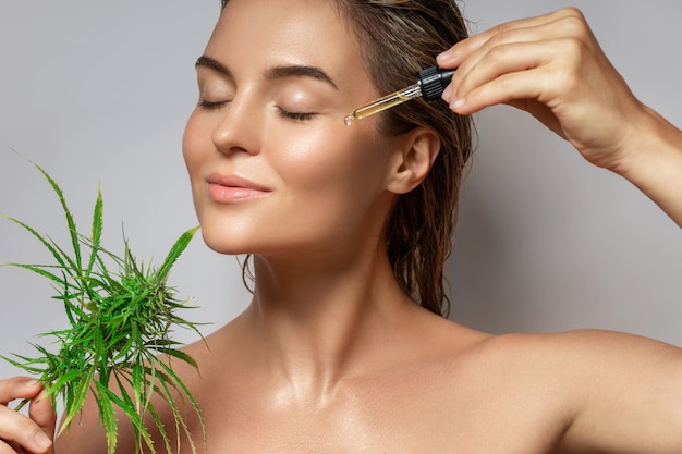 Concepto de cosméticos de CBD. Bella mujer con una hoja de cannabis sobre fondo gris