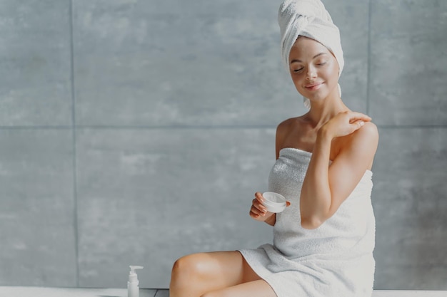 Concepto de cosmetología y spa de belleza para personas Mujer europea joven relajada que pone crema corporal toca el hombro suavemente envuelto en una toalla suave blanca cierra los ojos con poses de placer contra la pared gris