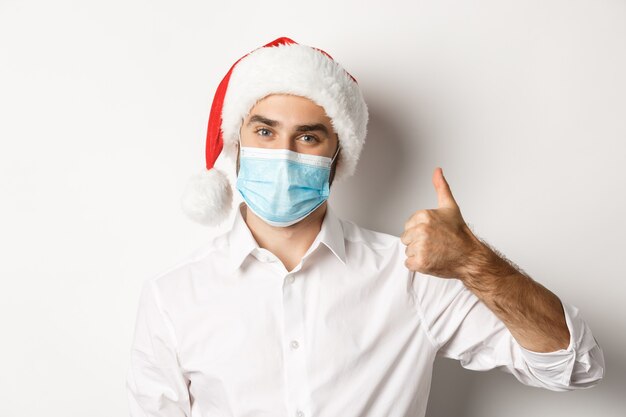 Concepto de covid-19, distanciamiento social y vacaciones de invierno. Hombre satisfecho con mascarilla y gorro de Papá Noel mostrando el pulgar hacia arriba, celebrando la Navidad con medidas preventivas, fondo blanco.