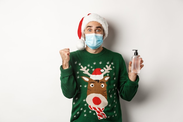 Concepto de covid-19 y vacaciones navideñas. Hombre alegre en mascarilla mostrando buen desinfectante de manos, vistiendo gorro de Papá Noel, de pie sobre fondo blanco.