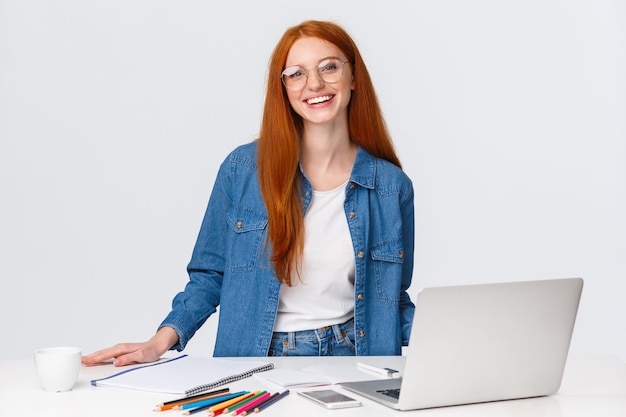 Concepto de creatividad, estilo de vida y educación. Linda chica pelirroja carismática con gafas de pie cerca de la mesa y la computadora portátil, prepara lápices de colores y personal para dibujar, aprender arte o diseñar cursos en línea