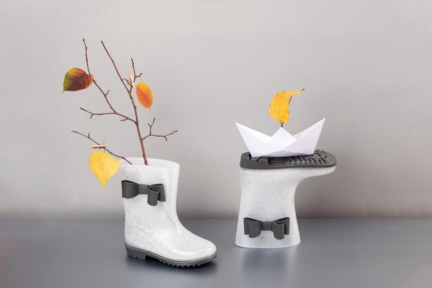 Concepto creativo de otoño Botas de lluvia de goma blanca brillante con arcos ramas de un árbol con hojas de otoño y barco de papel sobre fondo gris