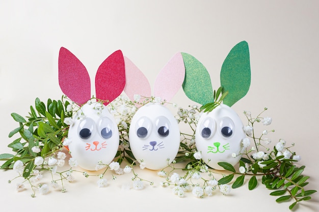 Concepto creativo de pascua: huevos en forma de conejitos con orejas de papel, ojos decorativos y bozales pintados. Alrededor - ramas con hojas verdes y pequeñas flores blancas. Copyspace