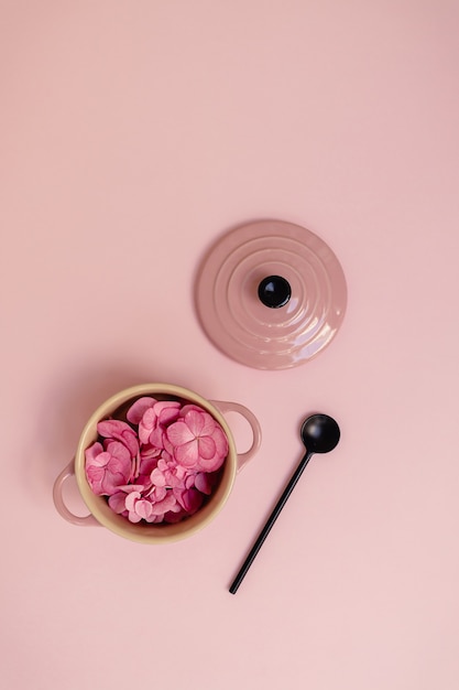 Concepto creativo con pétalos de flores de hortensias de primavera en una cacerola junto a una cuchara sobre fondo rosa pastel. Endecha plana de naturaleza mínima.