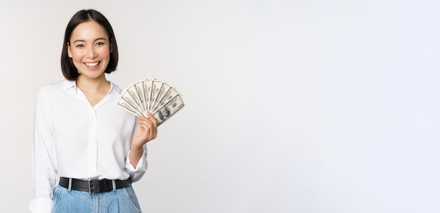Concepto de crédito y préstamo Mujer asiática joven sonriente sosteniendo dólares en efectivo y mirando feliz el fondo blanco de la cámara