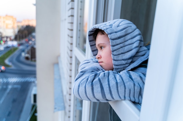 Concepto de cuarentena por coronavirus. Niño vestido con chaqueta con capucha durante el virus de la gripe, mirando por la ventana. COVID-19: autoaislamiento. Muchacho adolescente obligado a quedarse en casa.