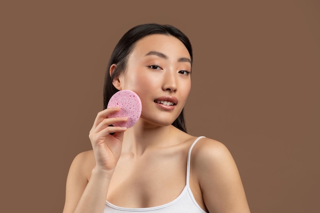 Concepto de cuidado de la piel bonita dama asiática sosteniendo una esponja cosmética lista para la limpieza facial posando sobre
